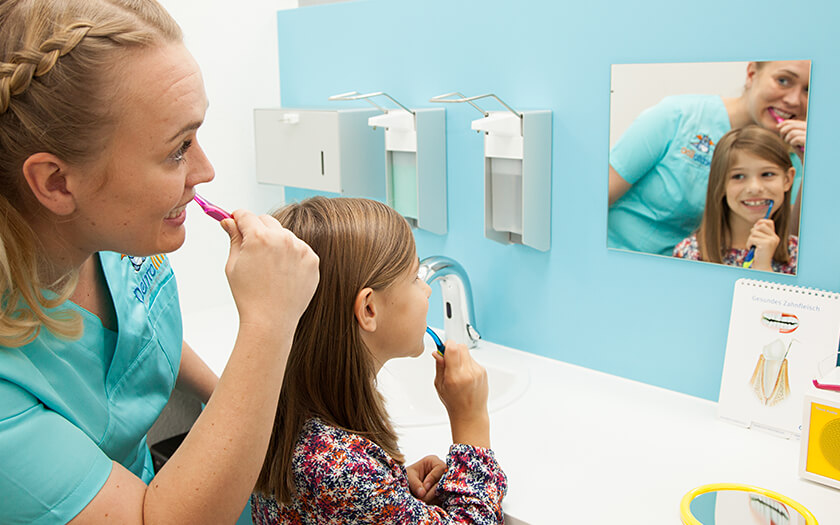 Zahnarzthelferin zeigt Mädchen im Spiegel bei Dentolino Ulm, wie man sich die Zähne putzt.